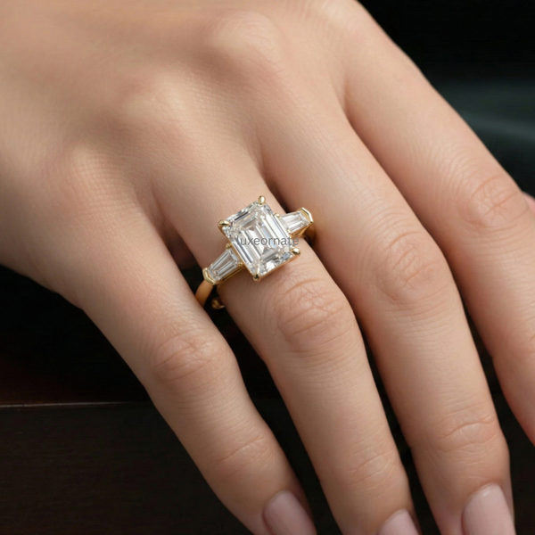 Emerald cut diamond engagement ring in yellow gold on hand, three stone ring with trapezoid side diamonds, lab grown diamond bridal ring, luxury proposal jewelry.